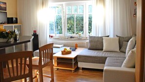 A cozy living room with a sofa, a wooden chair, a small table with fruit, and a window with curtains. - Hus Mattgoot (St. Peter-Ording)