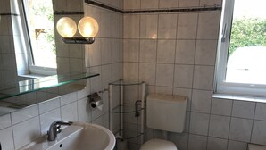 Et badeværelse med en hvid vask, toilet og bidet. Der er en glashylde over vasken og et vindue på højre side.