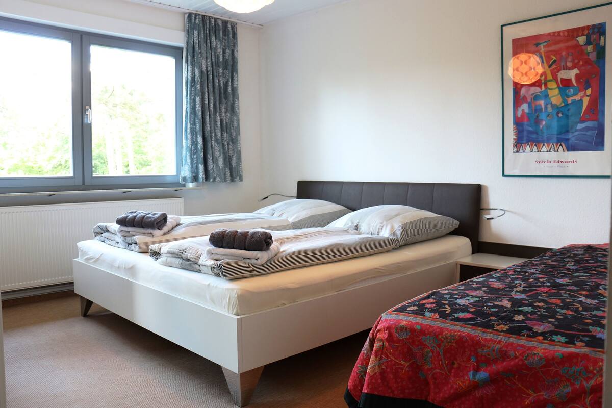 A double bed with a white headboard, a window with curtains, and a colorful artwork on the wall.