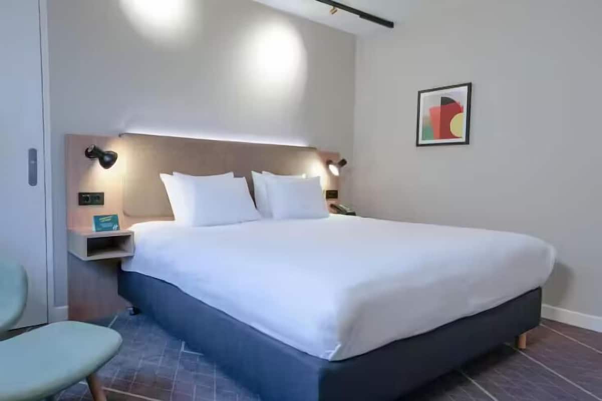 A modern hotel room with a large bed, a bedside table, a chair, and a wall-mounted artwork.