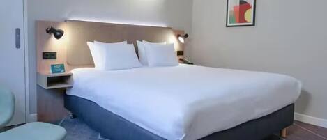 A modern hotel room with a large bed, a bedside table, a chair, and a wall-mounted artwork.