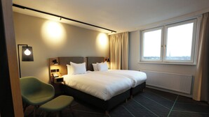 A hotel room with a large bed, two bedside tables, a green chair, and a window with a view. - Nordseka hotel The Hague Voorburg (Voorburg)