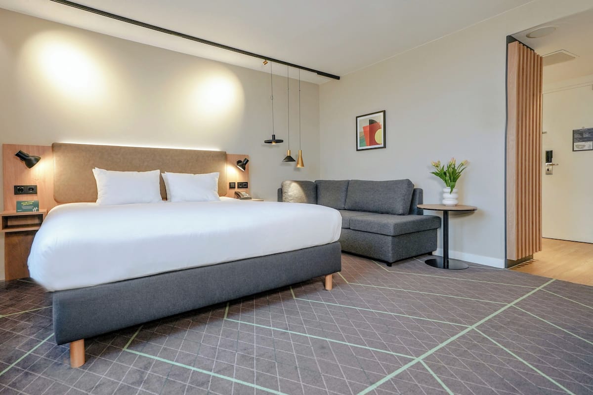 A modern hotel room with a large bed, a grey sofa, a small table with a vase of flowers, and a wooden headboard.