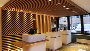 A modern reception area with a slatted wooden ceiling, two reception desks, and a large window providing a view of the street. - Nordseka hotel The Hague Voorburg (Voorburg)
