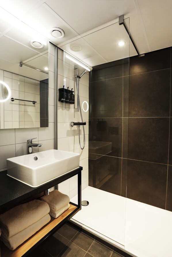 A modern bathroom with a white sink, dark tiled shower, and a mirror with lights. - Nordseka hotel The Hague Voorburg (Voorburg)