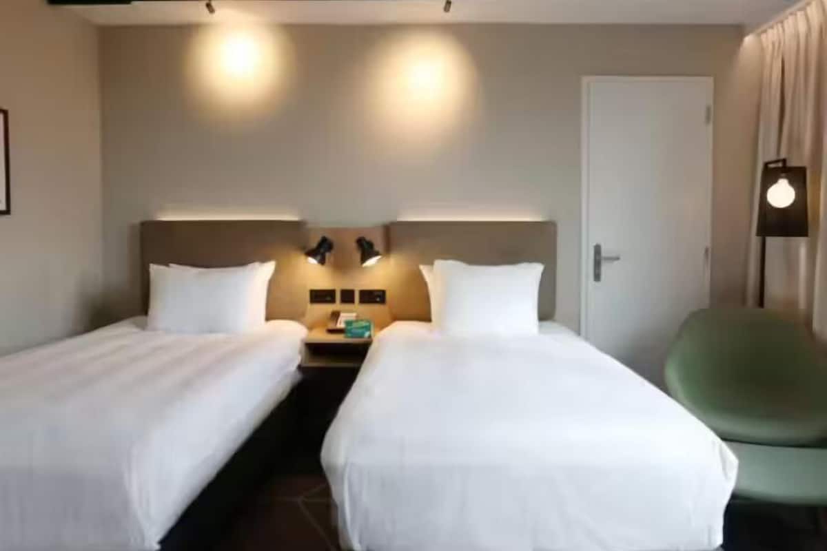 A hotel room with two single beds, a nightstand with a lamp, a green chair, and a door.