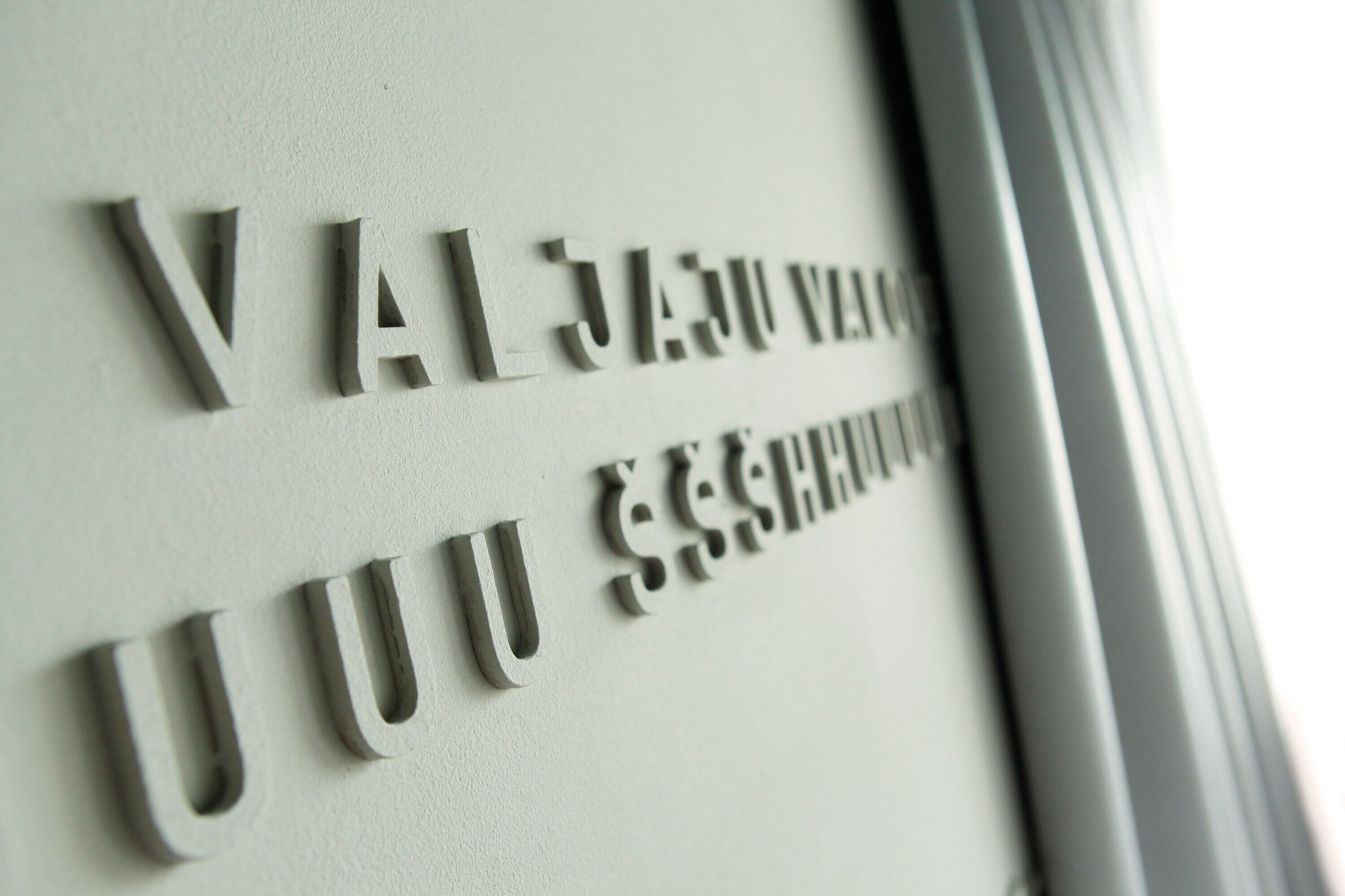 Primer plano de una pared con texto en relieve, probablemente un nombre o título, con una tipografía formal.
