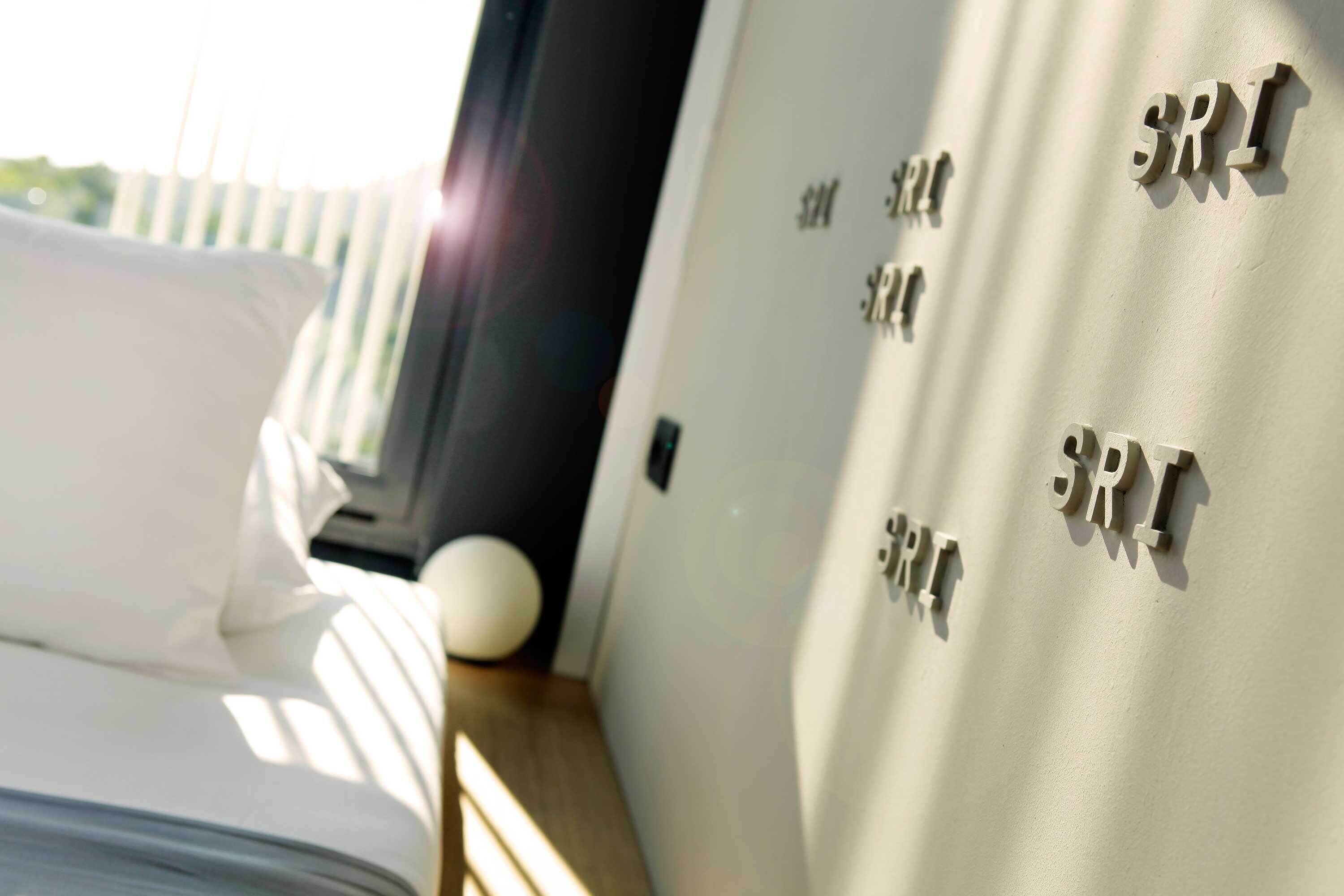 A hotel room with a bed, a pillow, a bedside table with a lamp, and a wall with 'SRI' letters.