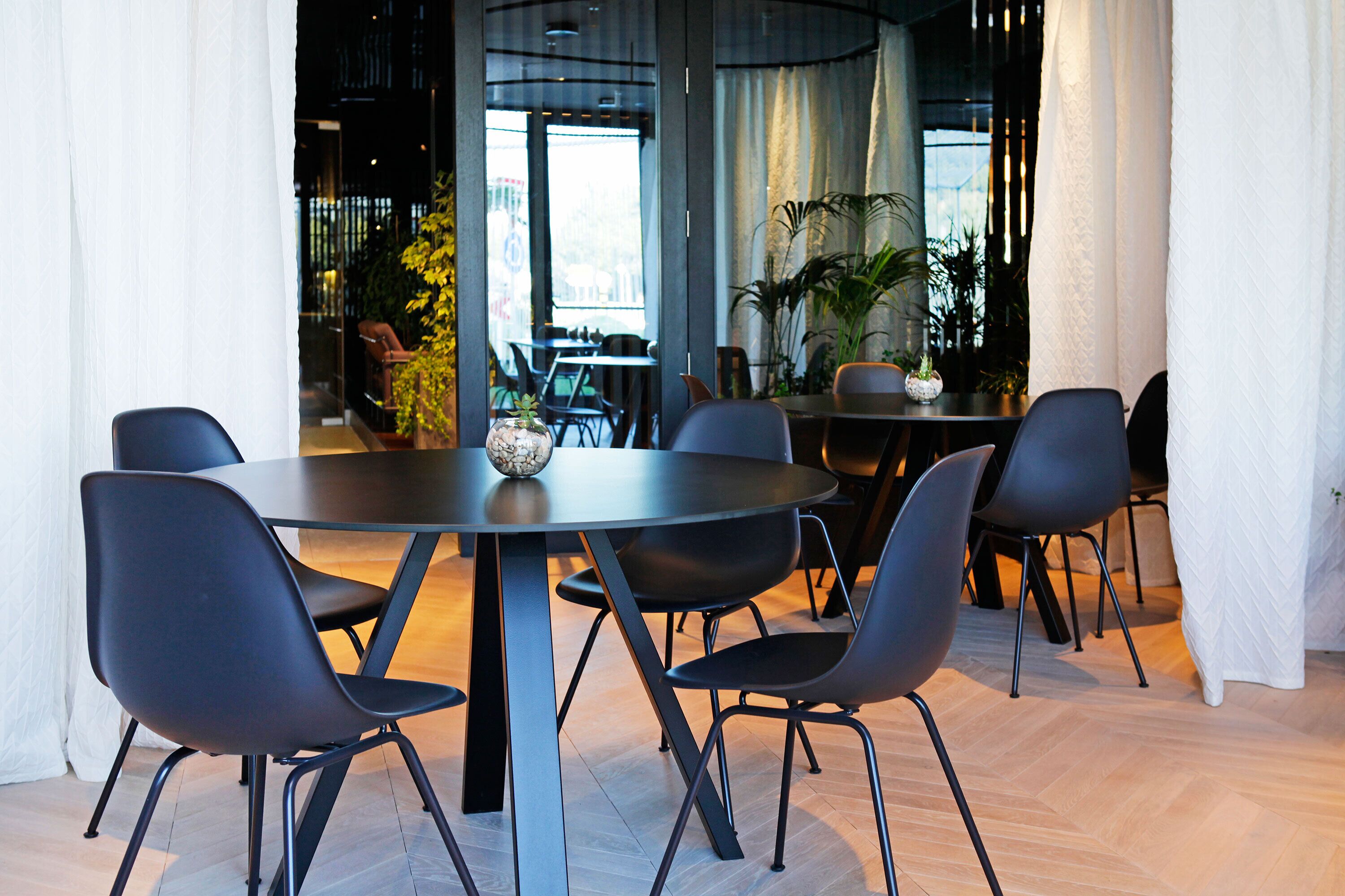 A modern dining area with a round table and black chairs, surrounded by large windows and indoor plants.
