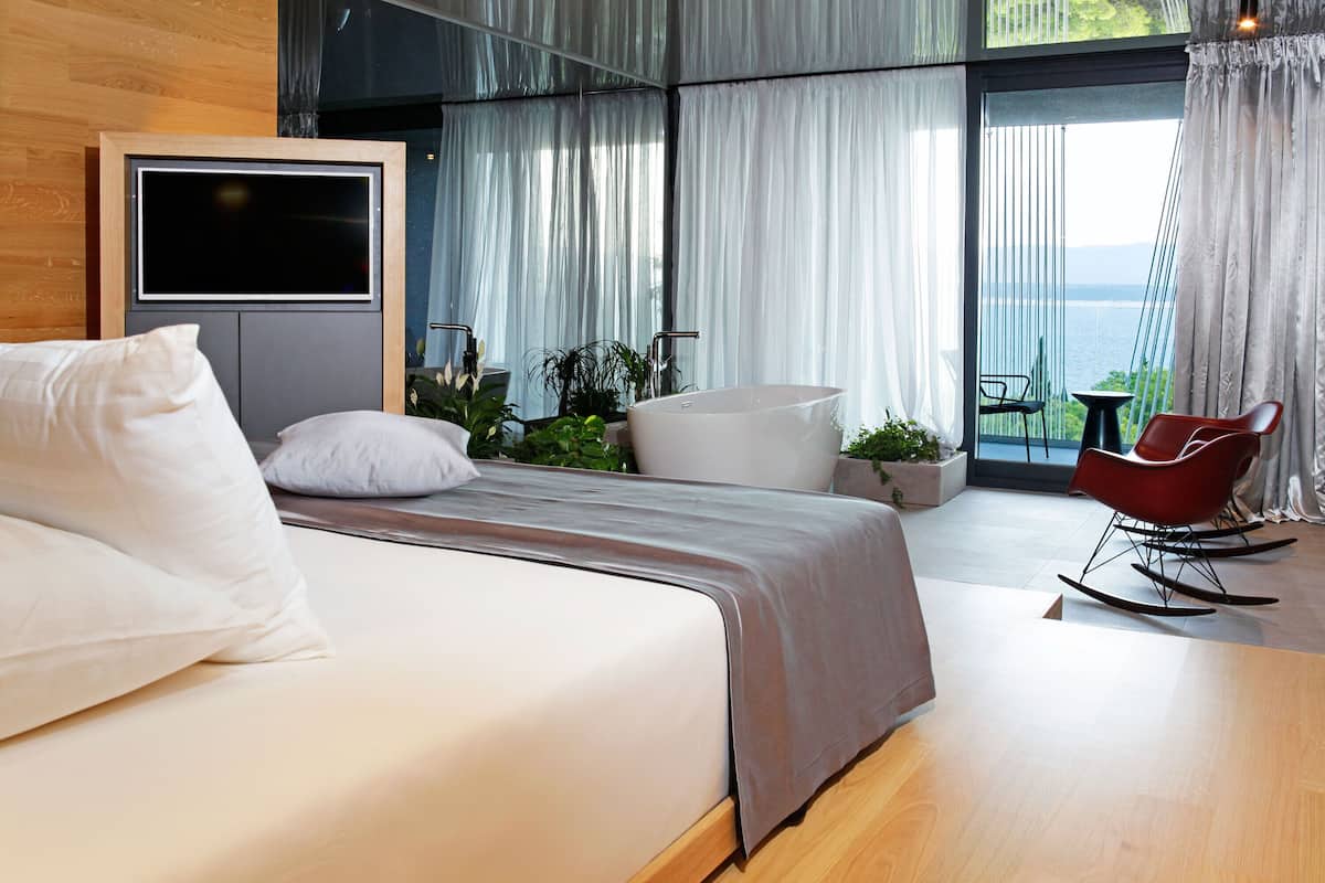 A modern bedroom with a large bed, a TV mounted on the wall, a balcony with a view of the sea, and a red rocking chair.