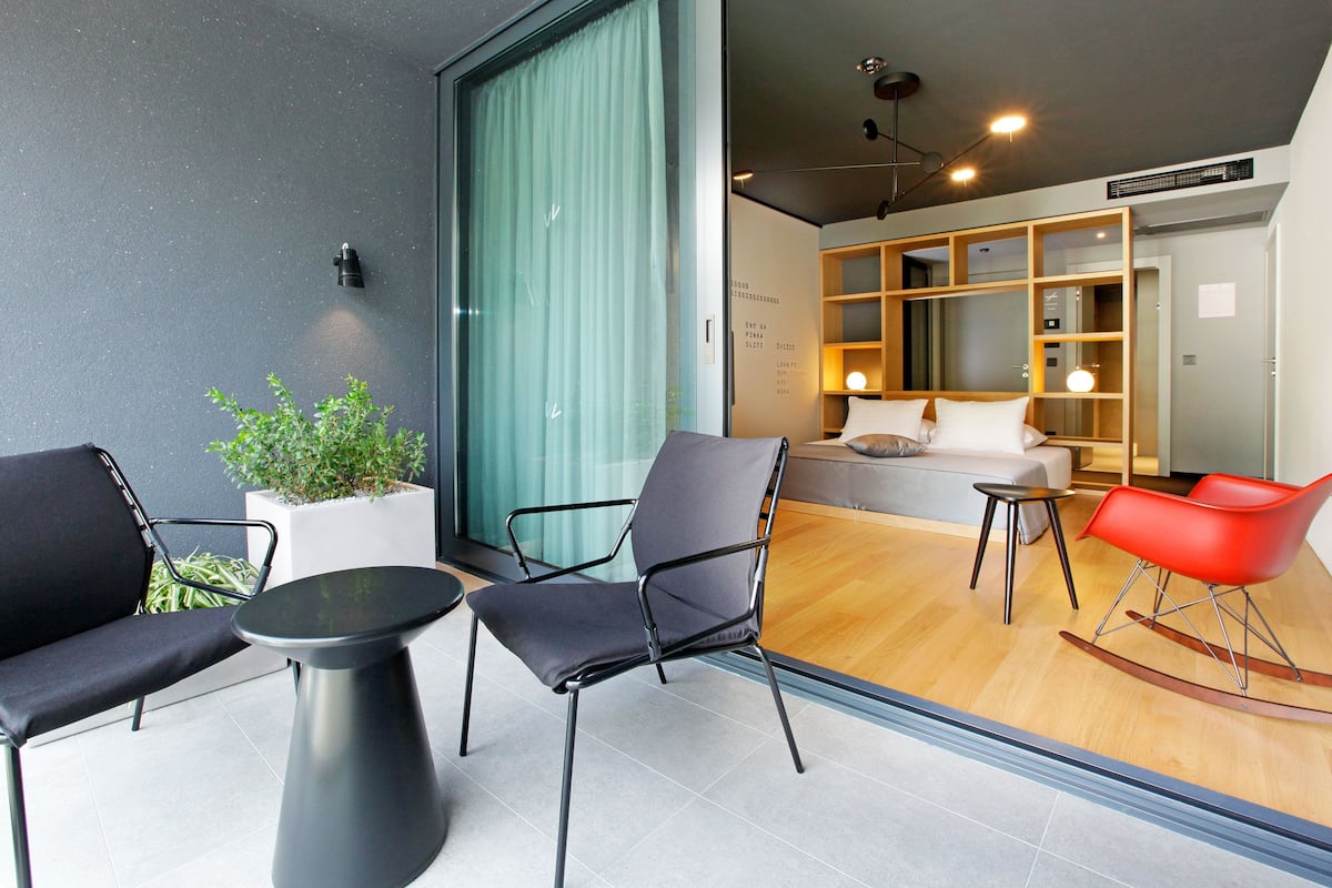 A modern hotel room with a sofa, chairs, a small table, a potted plant, and a wooden bookshelf.