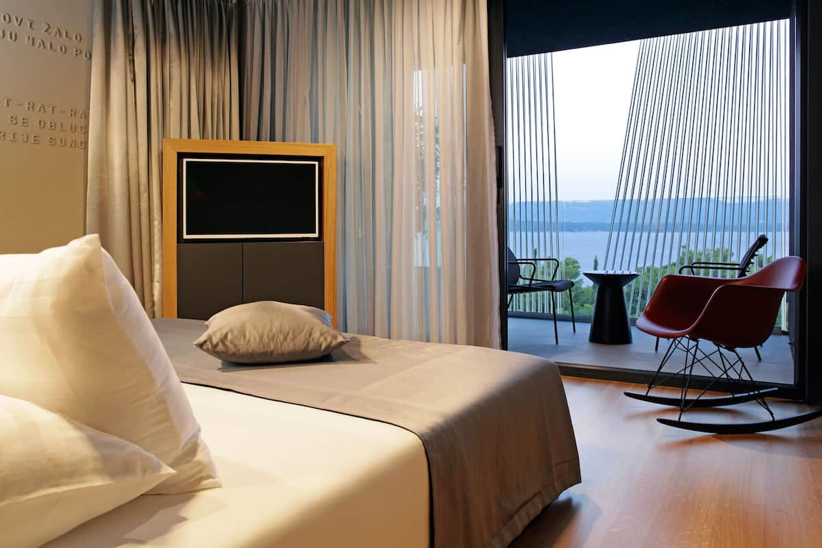 A hotel room with a bed, a TV, a balcony with a chair, and a view of the water.