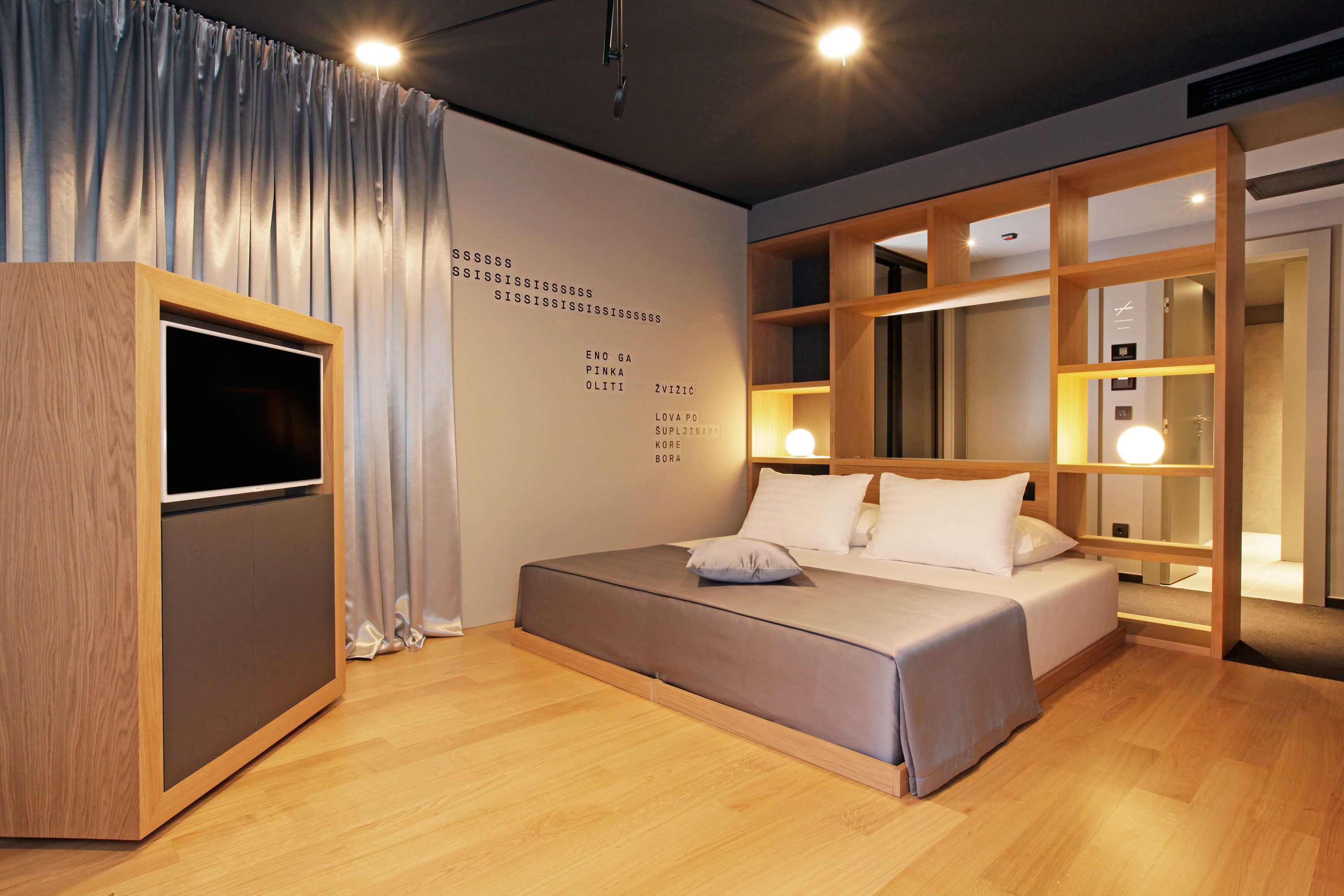 A modern hotel room with a bed, a TV, and a wooden shelf unit.
