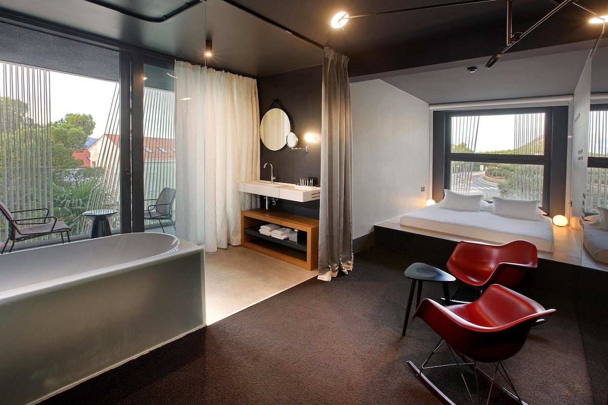 A modern bedroom with a large window, a bedside table, a red chair, and a bathtub.