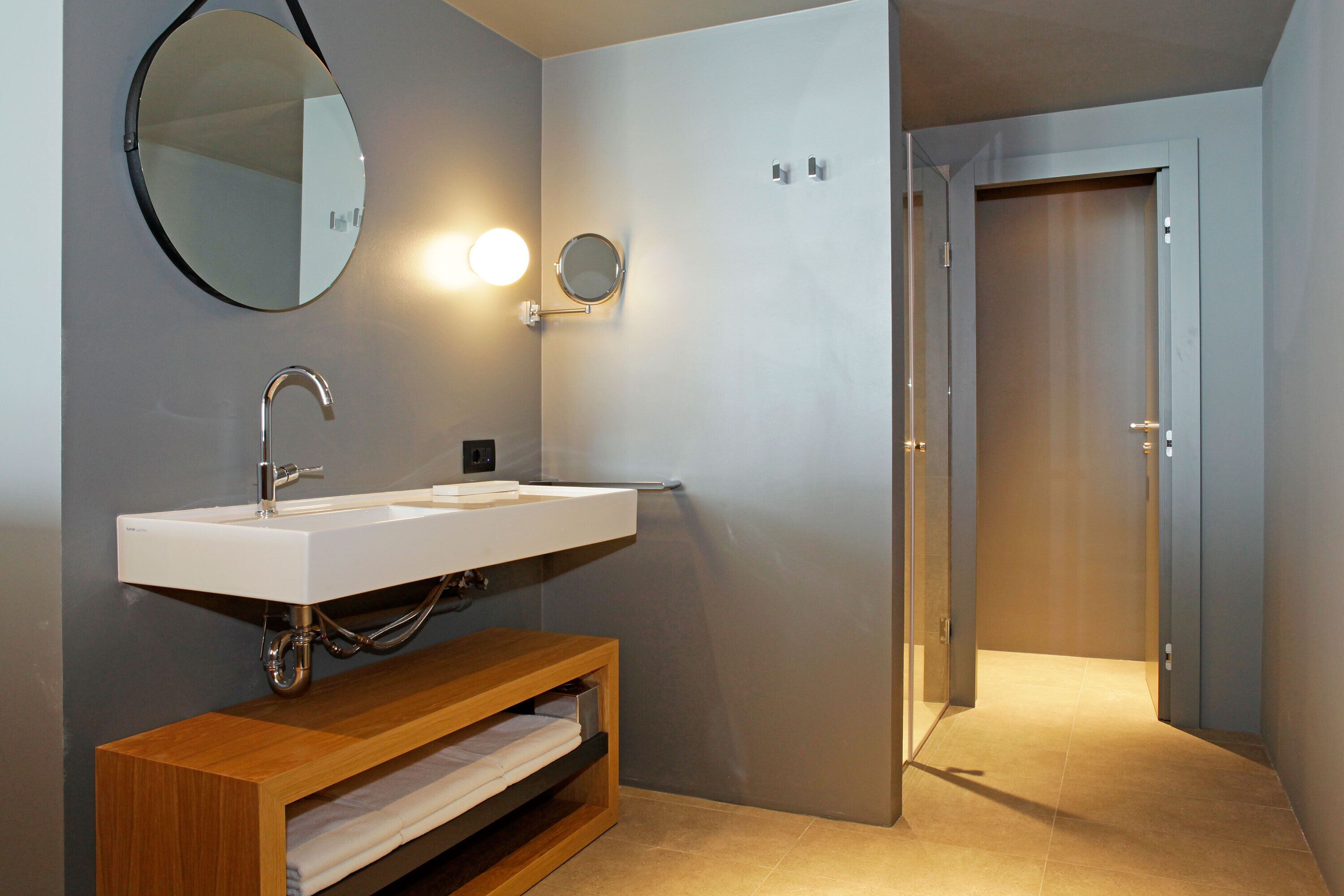 Un baño moderno con un lavamanos blanco, un mueble de tocador de madera, un espejo redondo y una puerta que da a otra habitación.