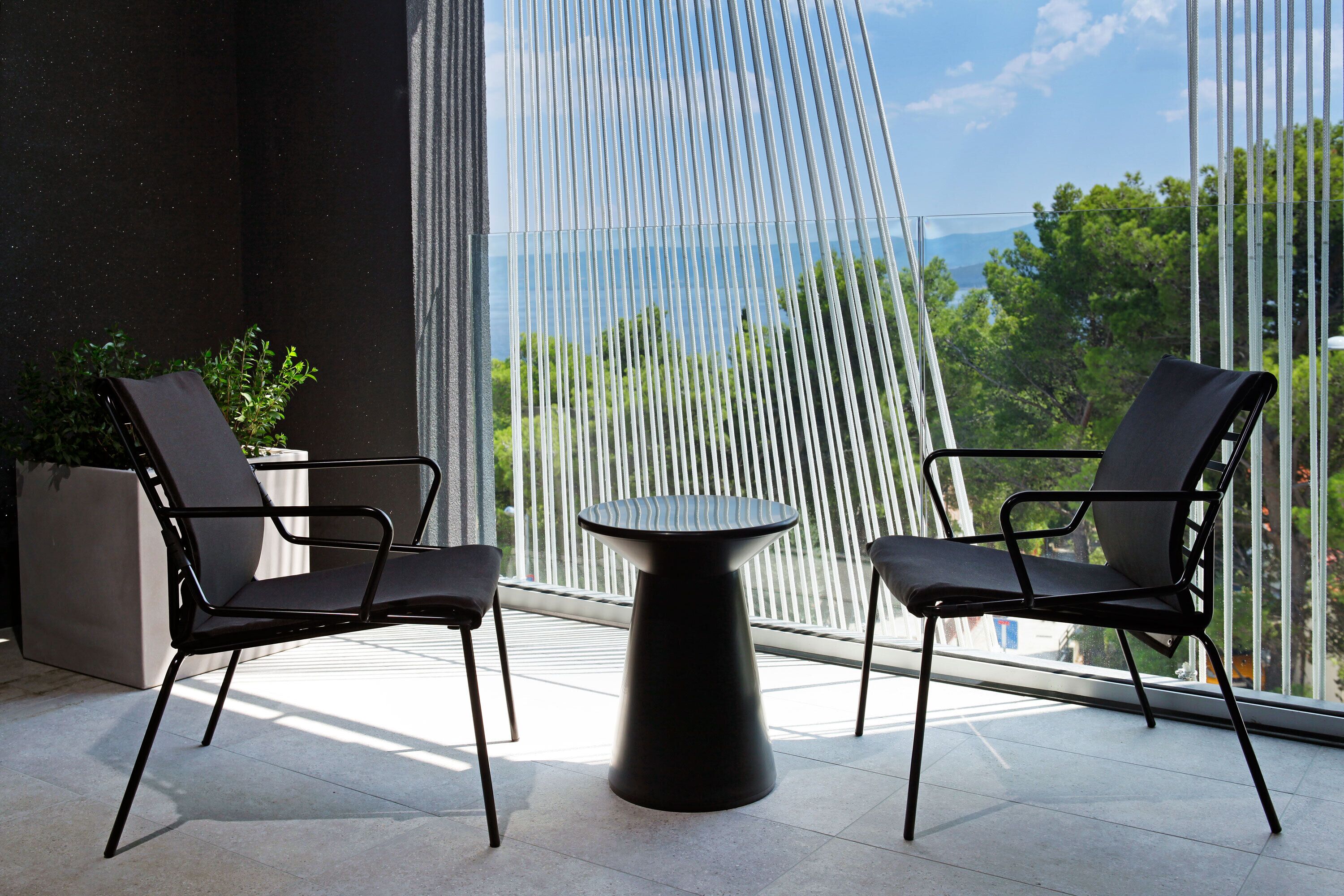 A modern outdoor seating area with two chairs and a table, large windows, and a view of greenery and mountains.
