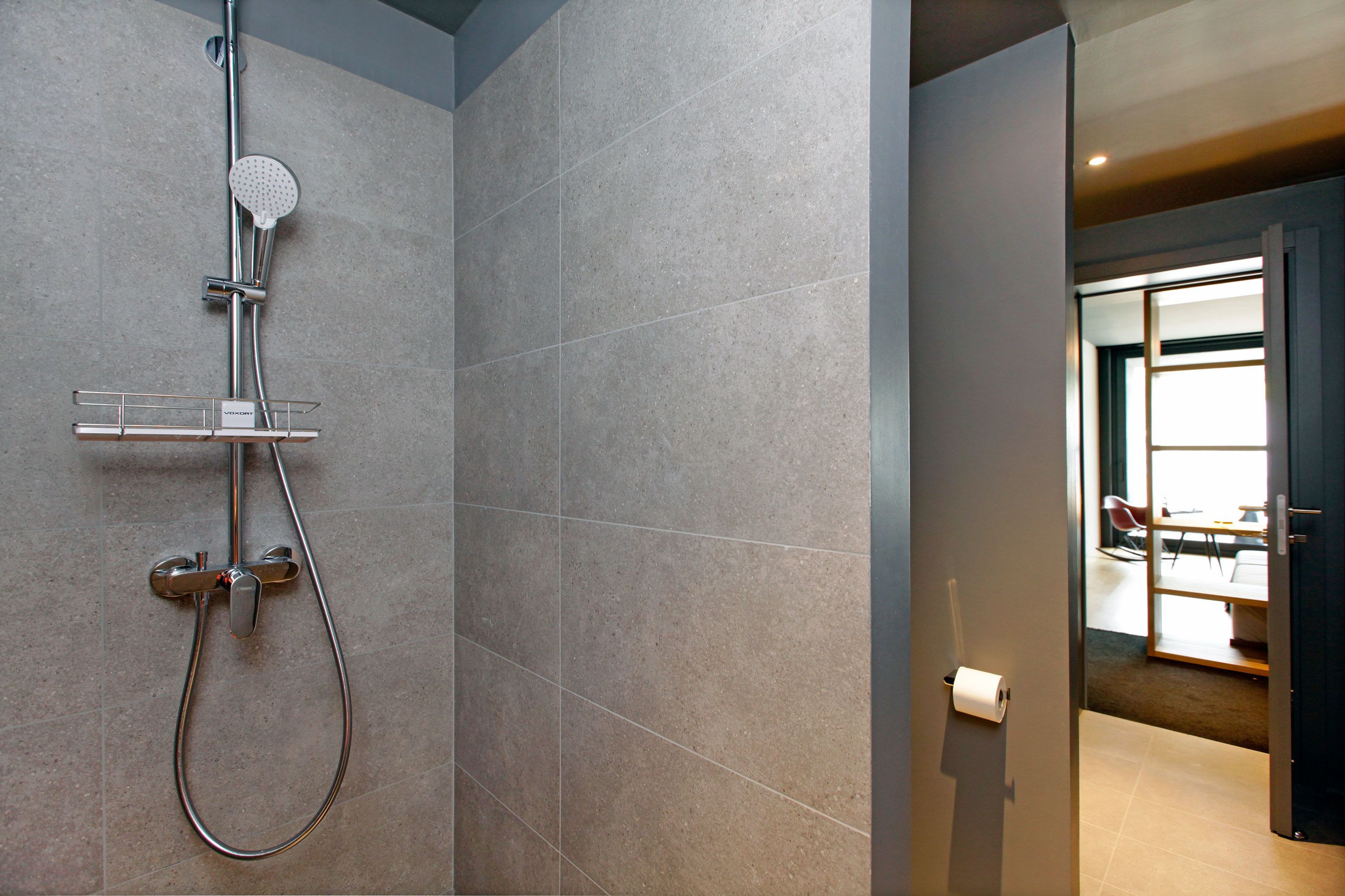 A modern bathroom with a shower, a toilet, and a visible door leading to a dining area.