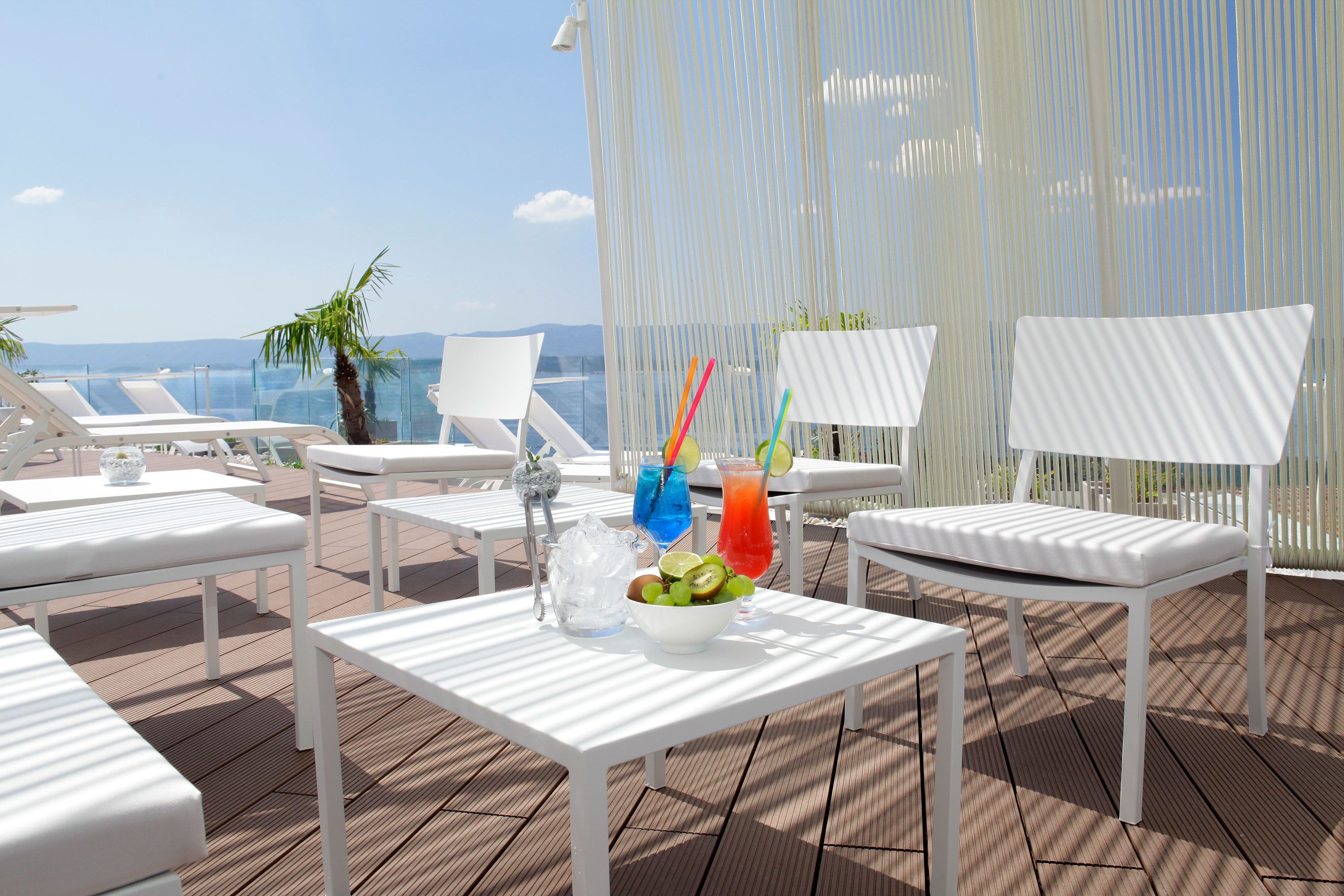 Un espacio al aire libre iluminado por el sol con sillones blancos y mesas, una mesa blanca con un cuenco de fruta y bebidas, y vistas al mar.