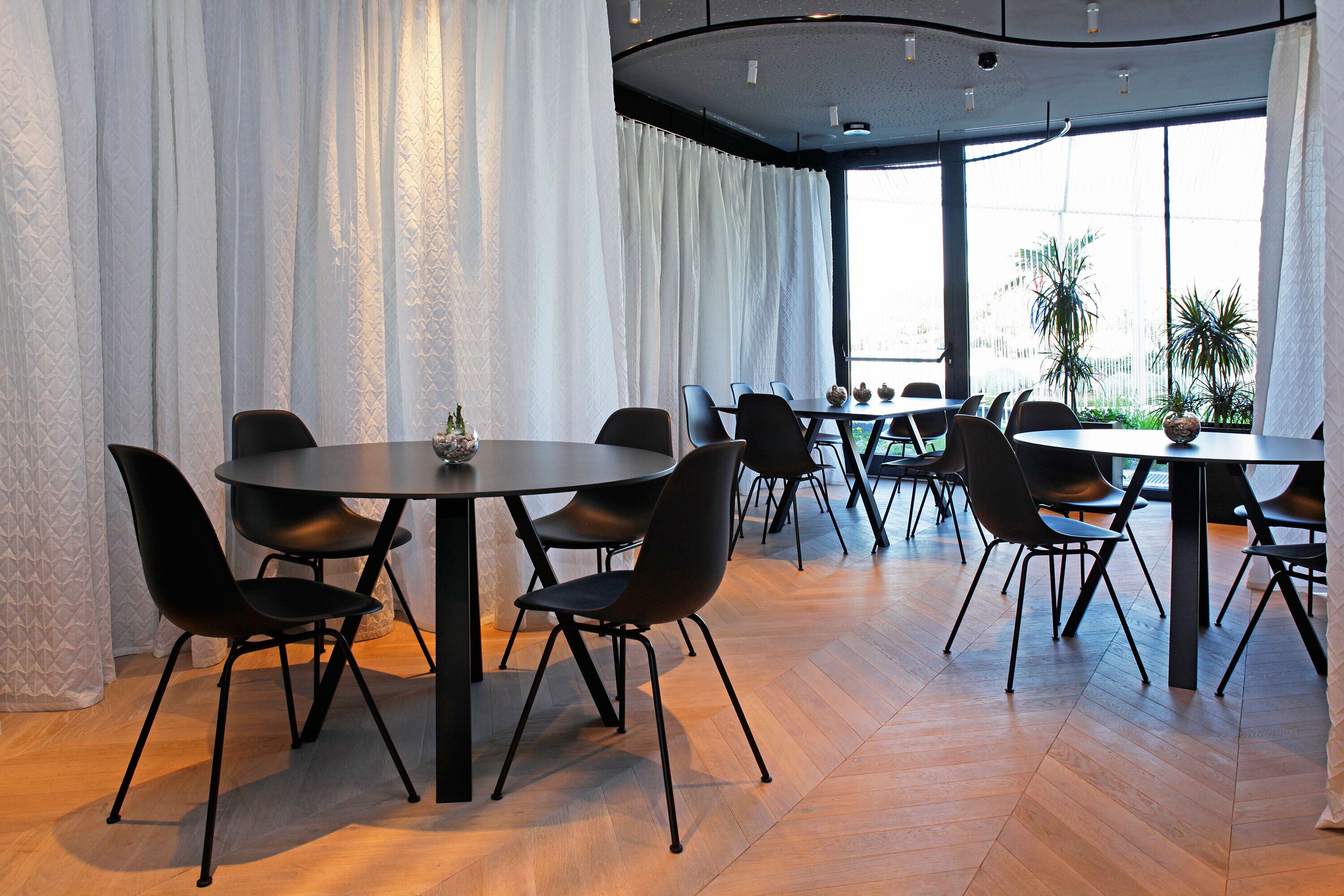 A modern dining area with round tables and black chairs, large windows with white curtains, and a view of palm trees outside.