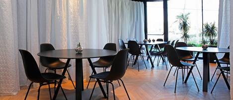 A modern dining area with round tables and black chairs, large windows with white curtains, and a view of palm trees outside.