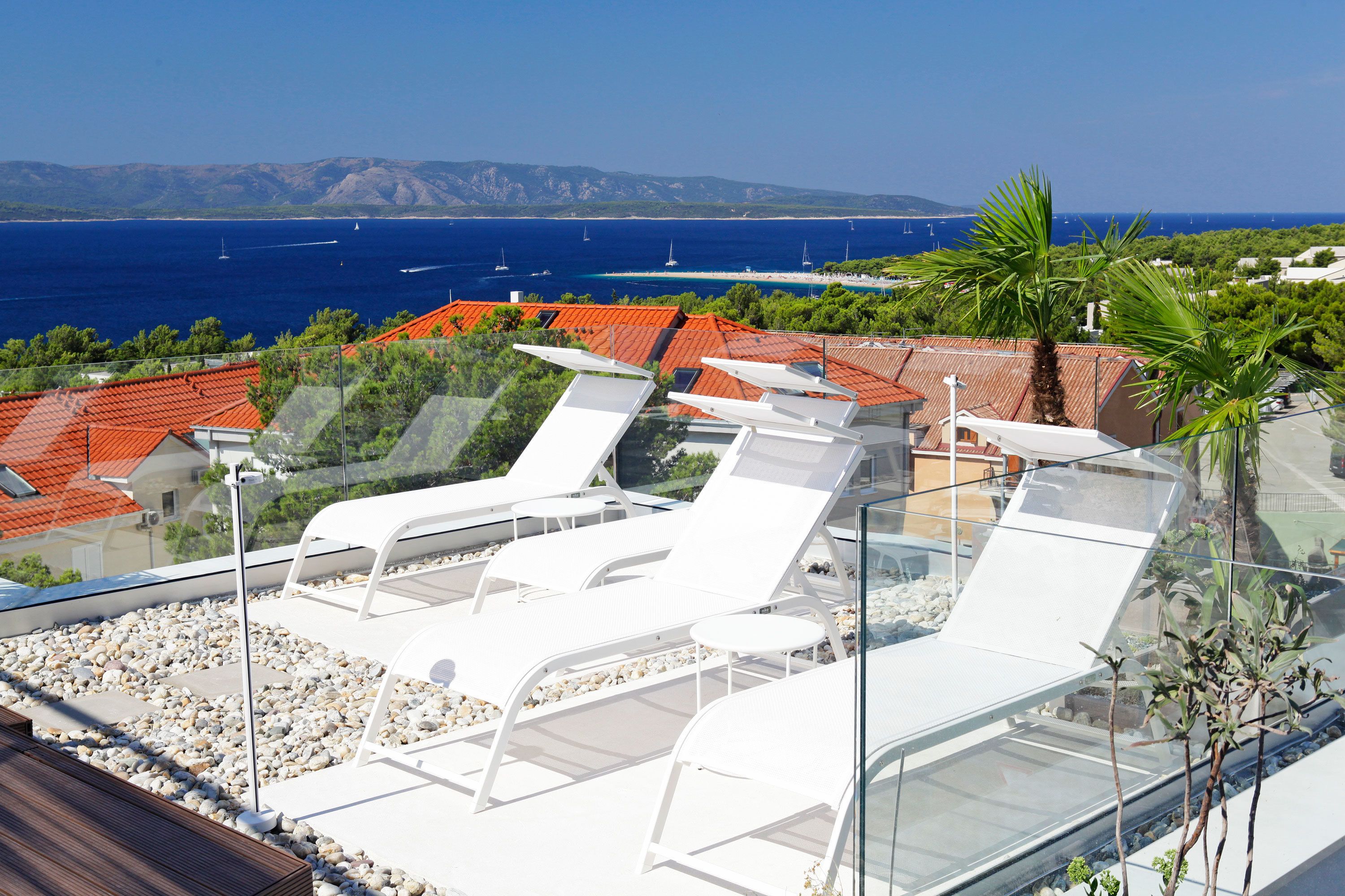 Une terrasse sur le toit, dotée de chaises longues blanches, d’une balustrade en verre et offrant une vue sur la mer et les montagnes lointaines.