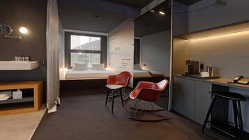 Une chambre d’hôtel moderne équipée d’un lit, d’un bureau avec un ordinateur, d’une chaise, d’une petite table et d’une étagère contenant des articles de toilette.