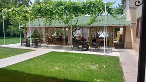 Un patio al aire libre con mesas y sillas, cubierto por una pérgola con cortinas blancas.