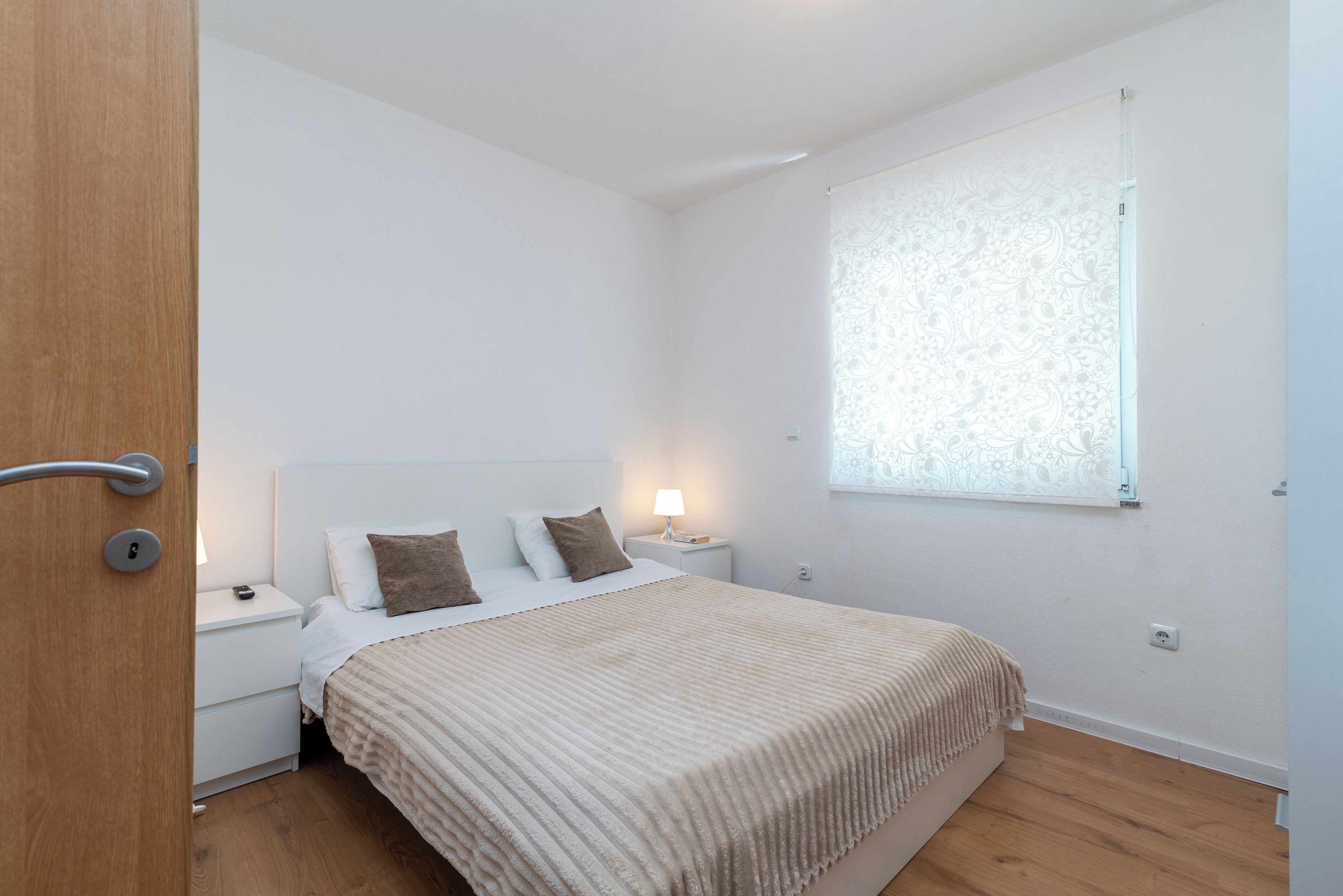 A bedroom with a bed, nightstands, a lamp, a window with patterned curtains, and a wooden door.