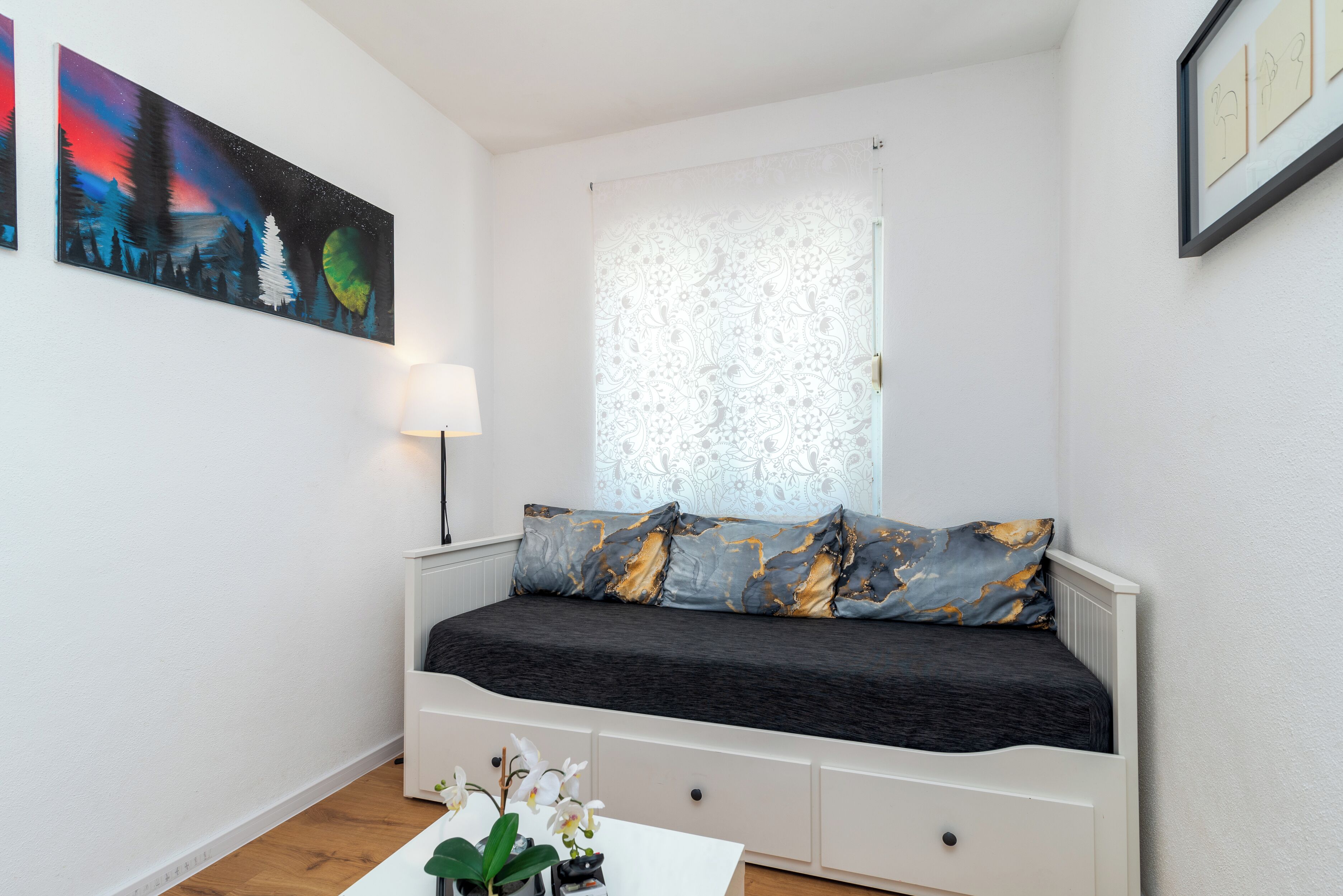 A bedroom with a bed, a window with patterned curtains, a bedside table with a lamp, and a painting on the wall.
