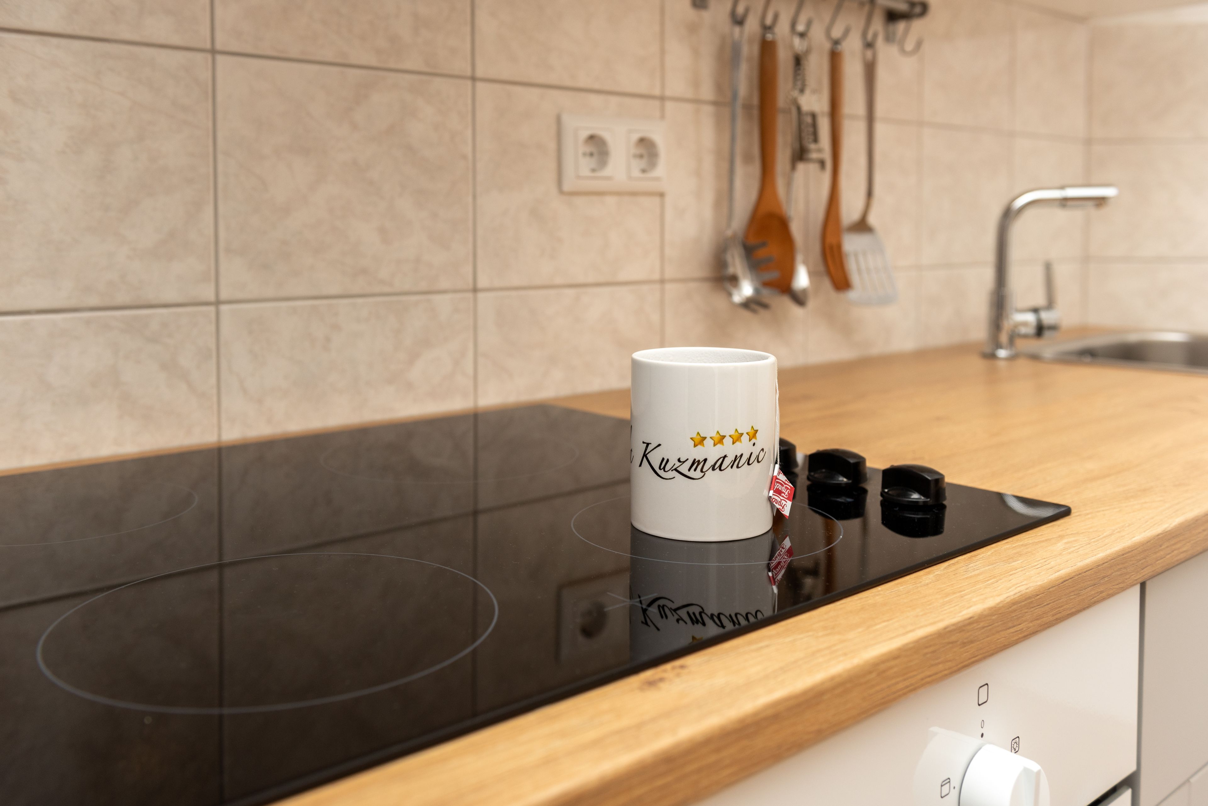 A kitchen with a black induction cooktop, a white mug labeled 'Kuzmanico' with three stars, and wooden utensils hanging on the wall.