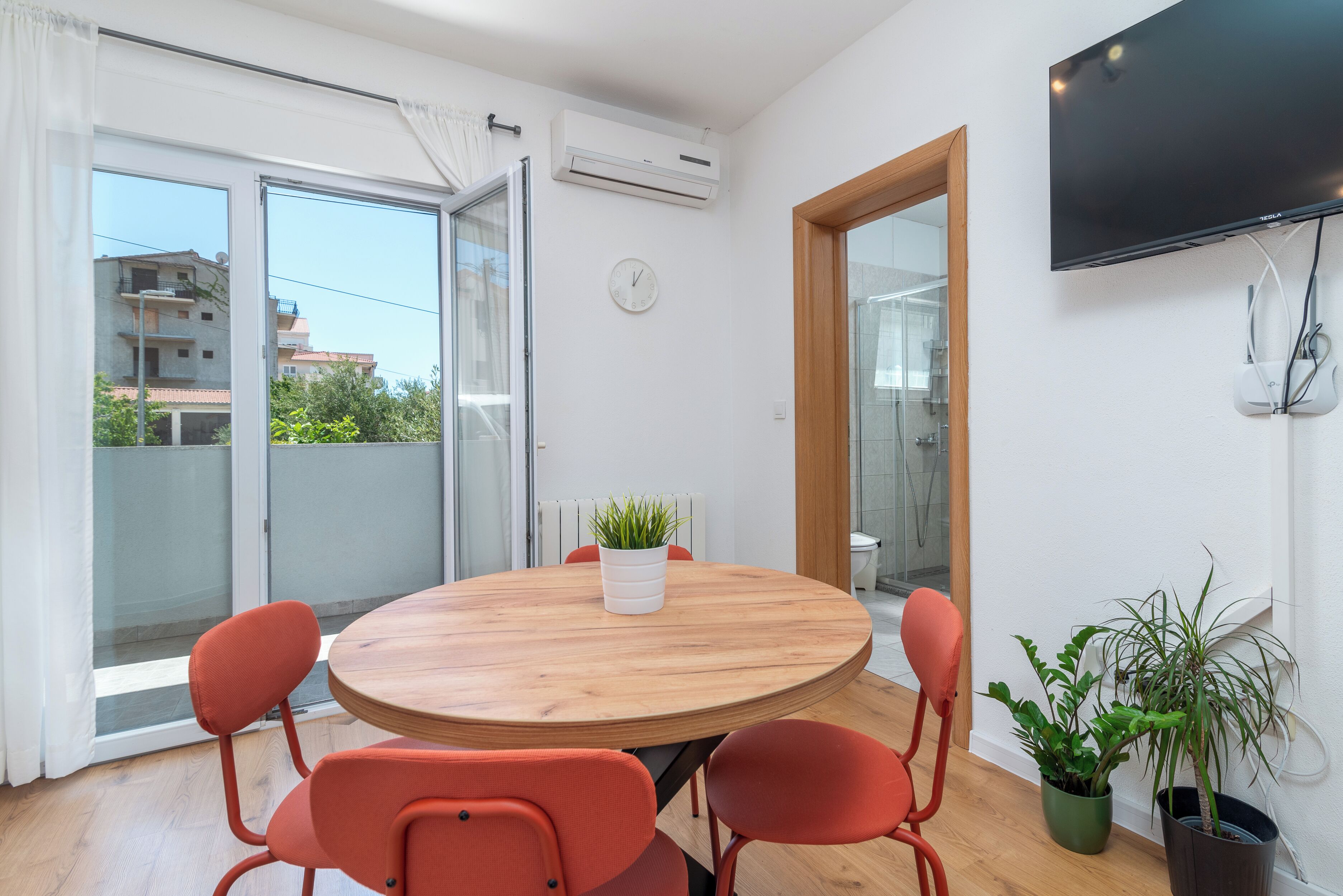 Une pièce avec une table ronde en bois, quatre chaises rouges, une plante en pot, un téléviseur fixé au mur et une porte-fenêtre coulissante donnant sur un espace extérieur.