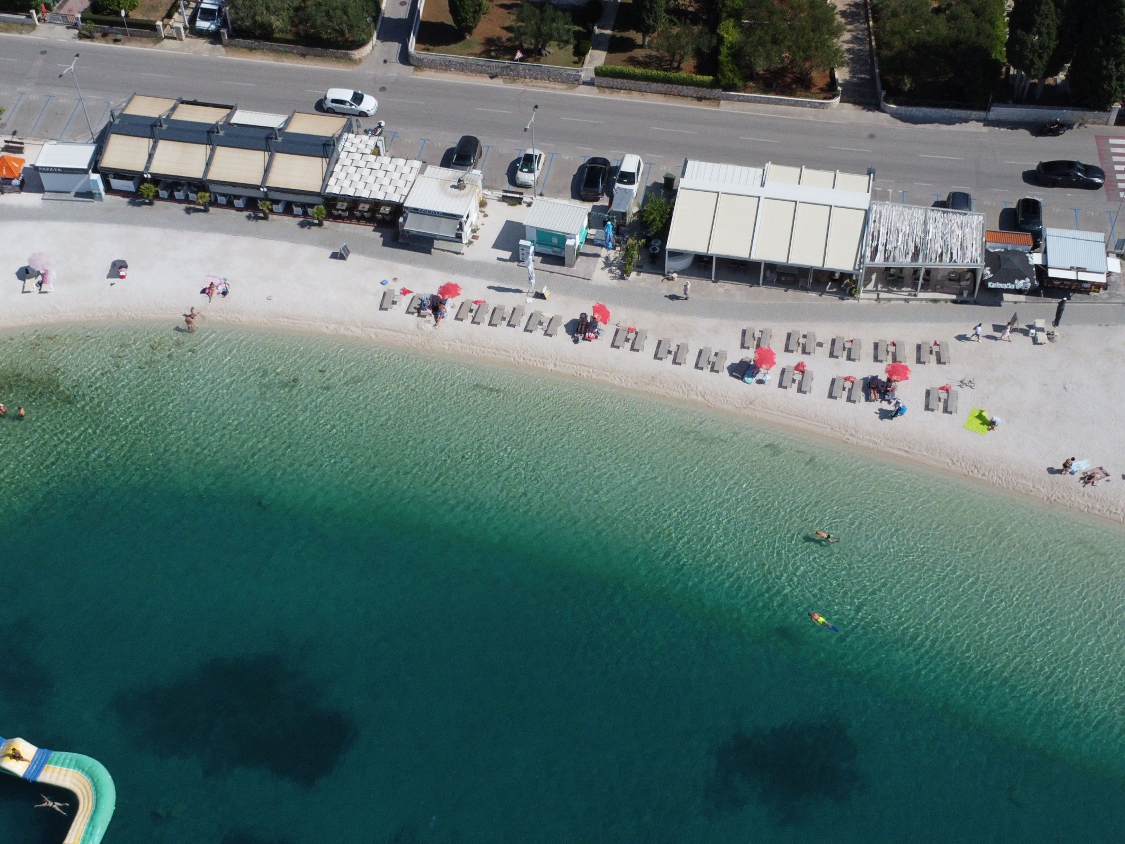 Eine Vogelperspektive zeigt einen Strand mit Liegestühlen und Sonnenschirmen, einen Parkplatz sowie Gebäude im Hintergrund.