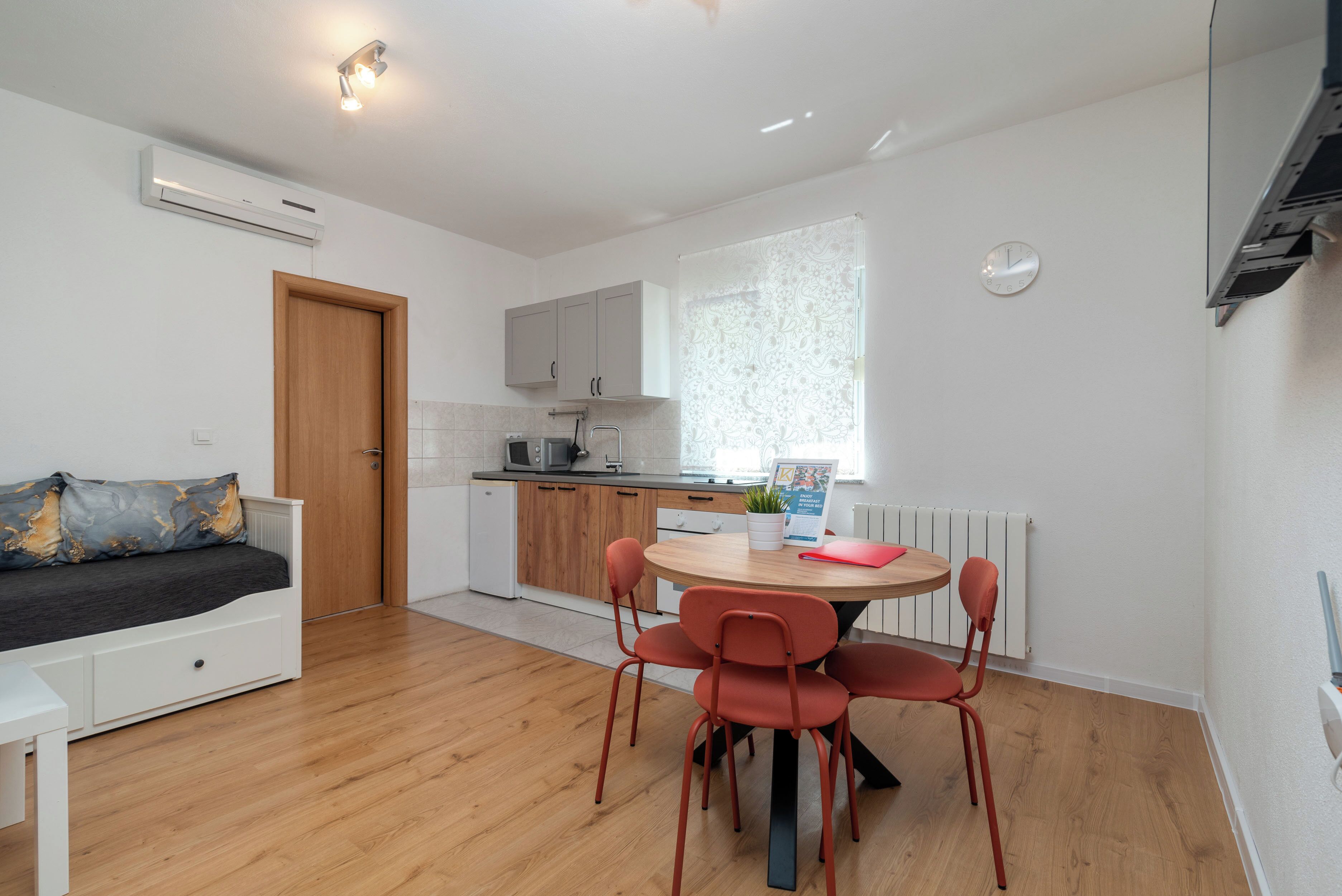 A modern kitchen with white cabinets, a wooden dining table with red chairs, a built-in oven, and a window with patterned curtains.