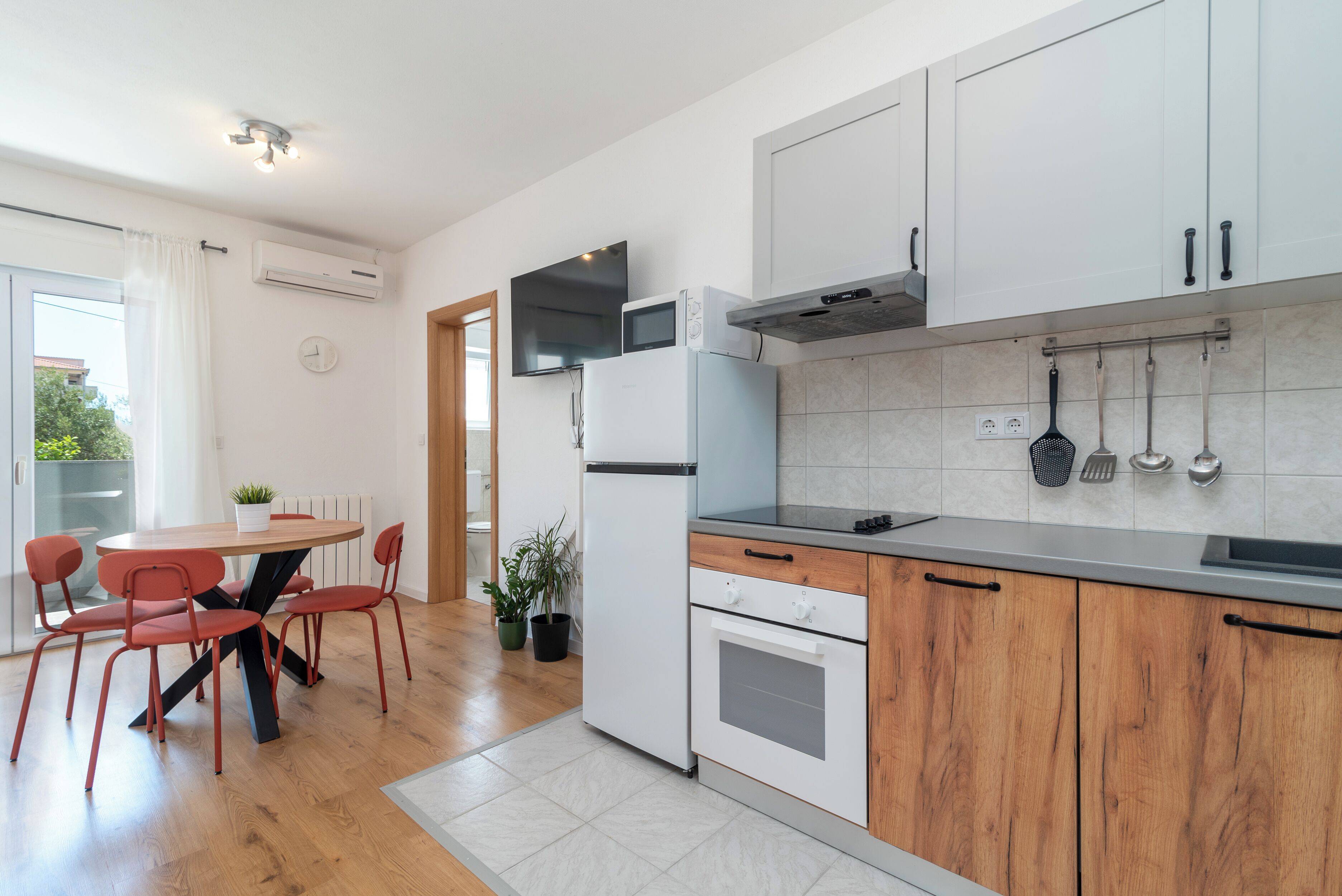 Una cocina moderna con armarios blancos, horno empotrado y zona de comedor con una mesa redonda y sillas rojas.