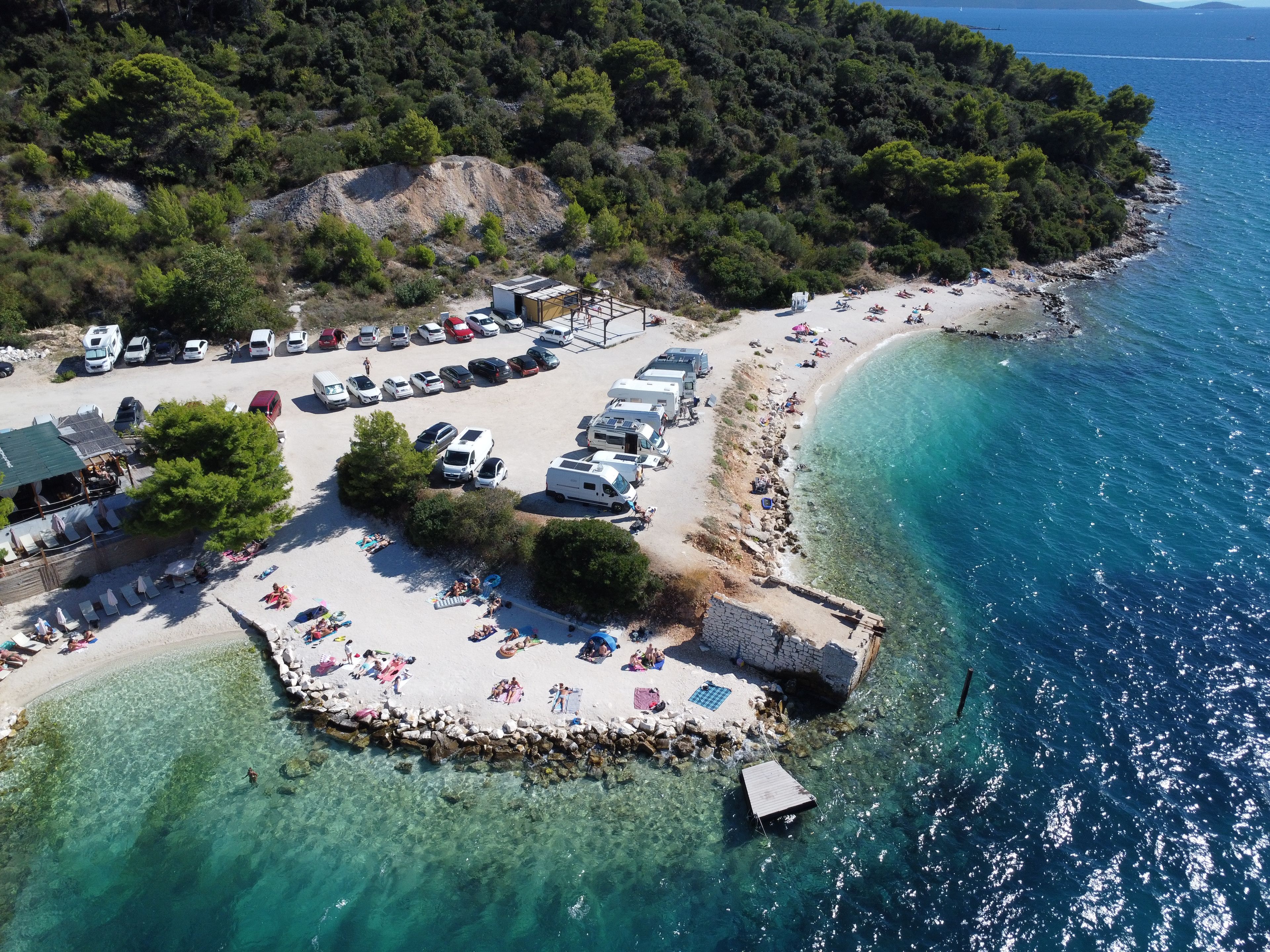An aerial view of a coastal area with a beach, parked vehicles, and a small dock.
