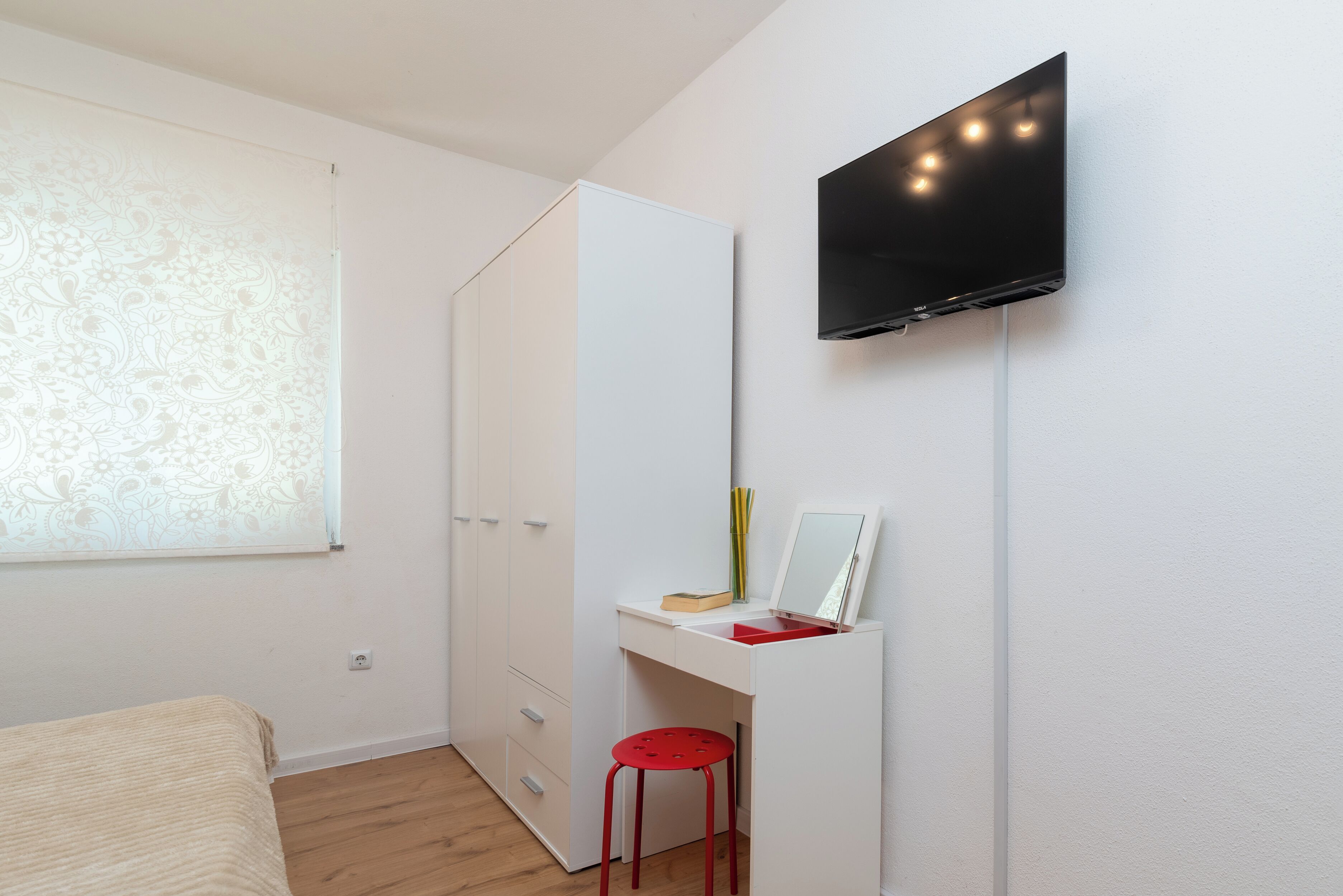 A bedroom with a bed, a desk with a mirror, a TV mounted on the wall, and a wardrobe.