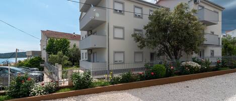 Edifício residencial de vários andares com varandas, entrada com calçamento de brita e um jardim cercado com flores.