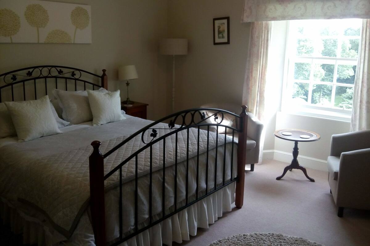 Quarto bem arrumado com cama, mesa de cabeceira, abajur, cadeira e janela com cortinas.