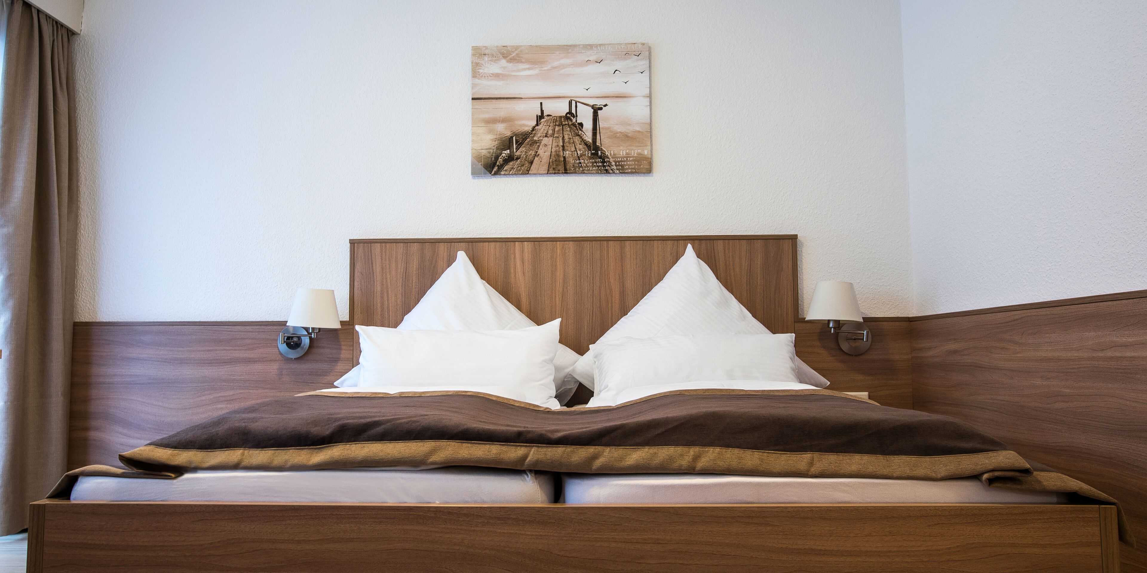 Una cama bien hecha con cabecera de madera, dos lámparas de noche y un cuadro enmarcado sobre ella.