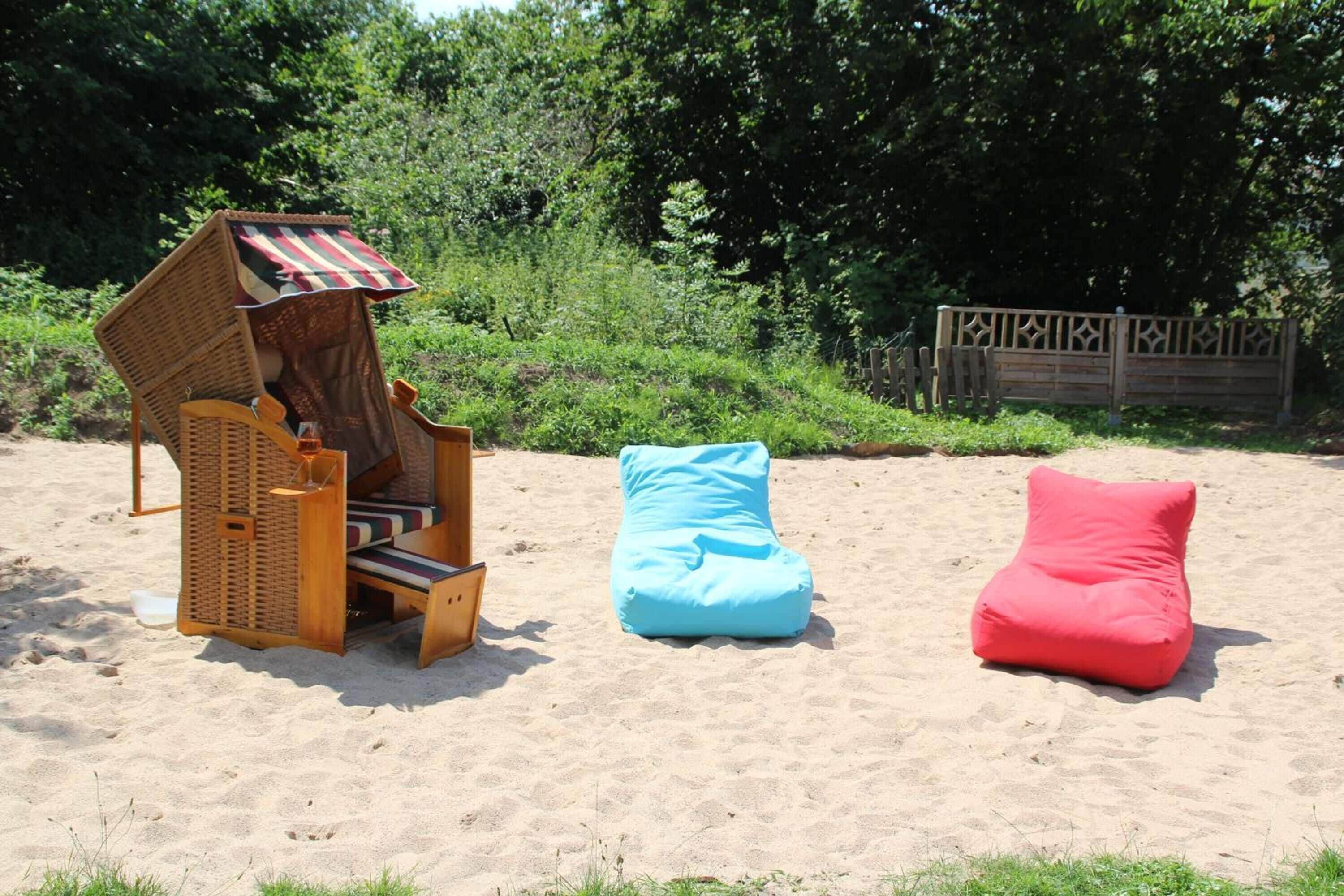 A beach setting with a wicker chair, a blue bean bag, and a red cushion on the sand.