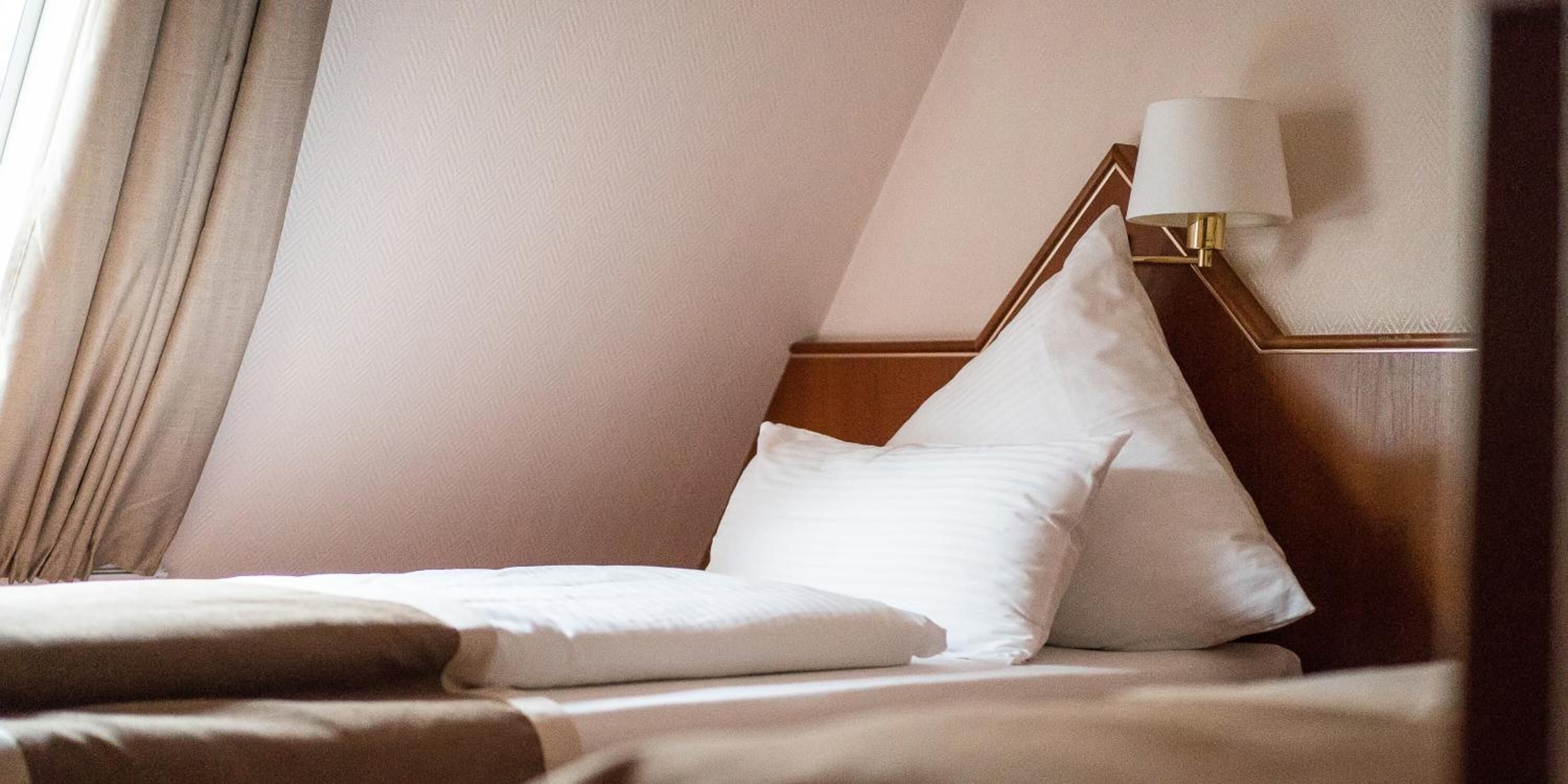 A neatly made bed with white linens and pillows, a wooden headboard, and a wall-mounted lamp with a white shade.