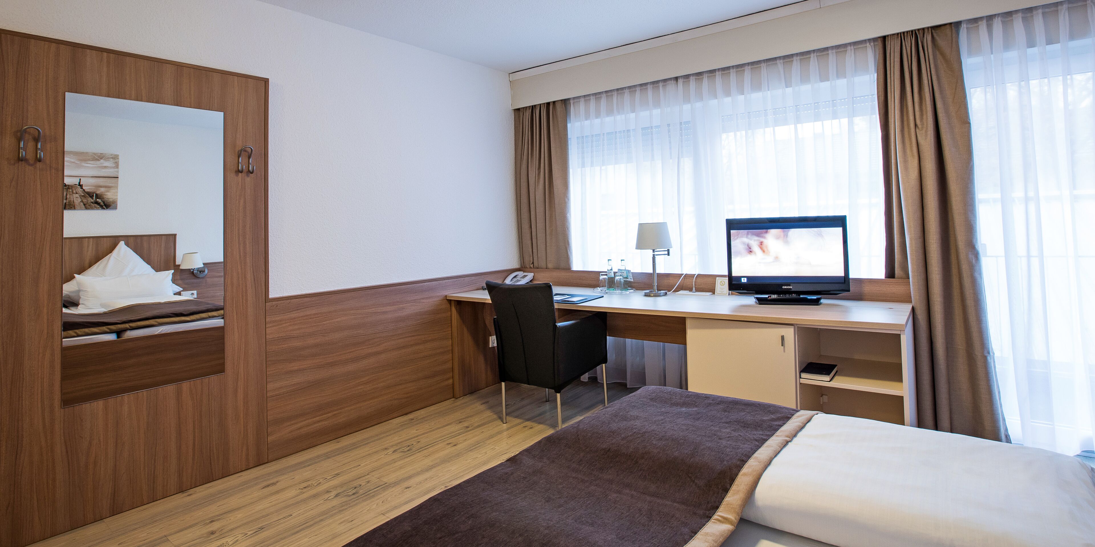 Ein Hotelzimmer mit einem Bett, einem Schreibtisch, einem Sessel, einem Fernseher und einem Vorhangfenster.