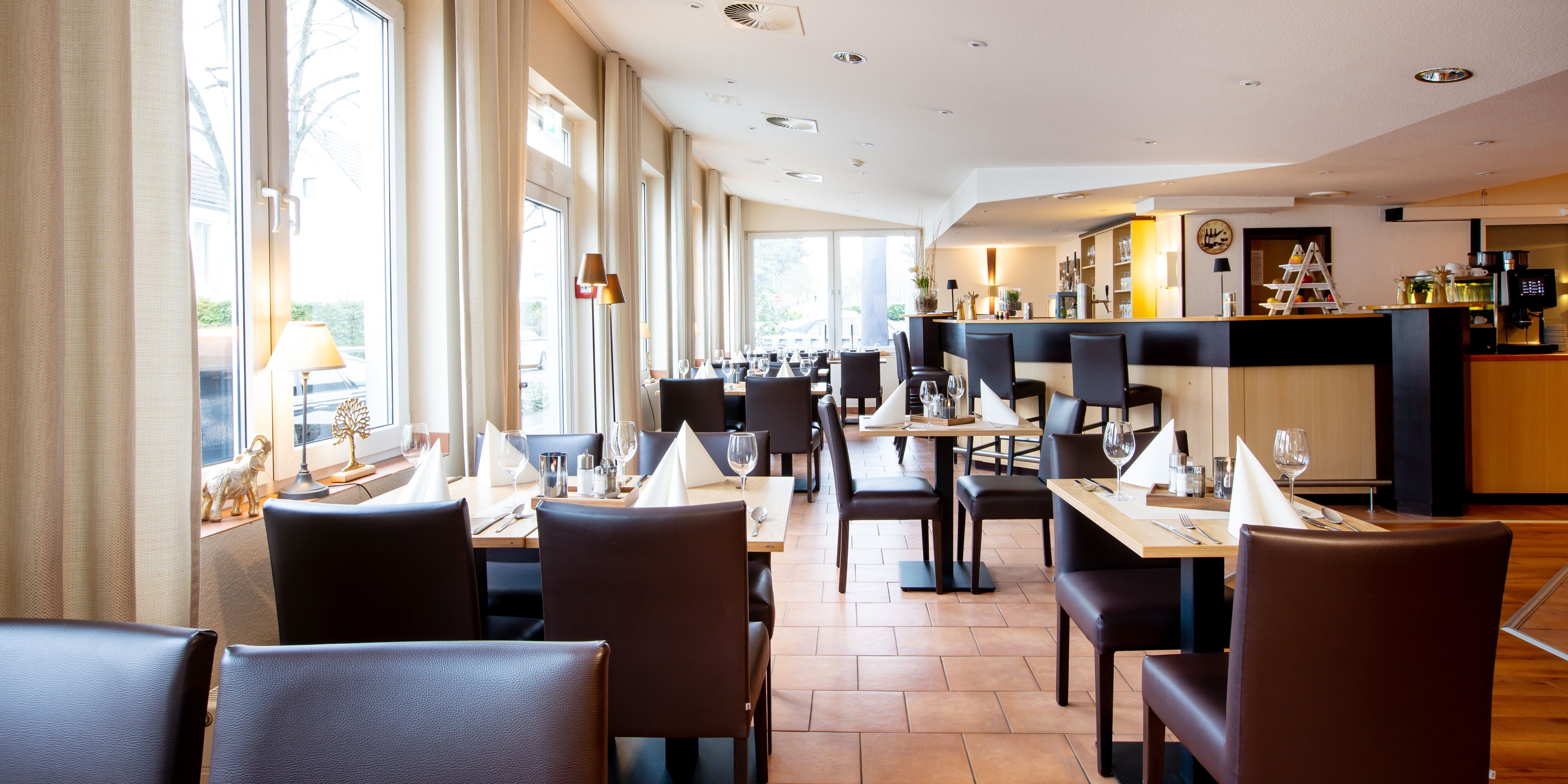 A restaurant with multiple tables and chairs, a bar area, and large windows allowing natural light.
