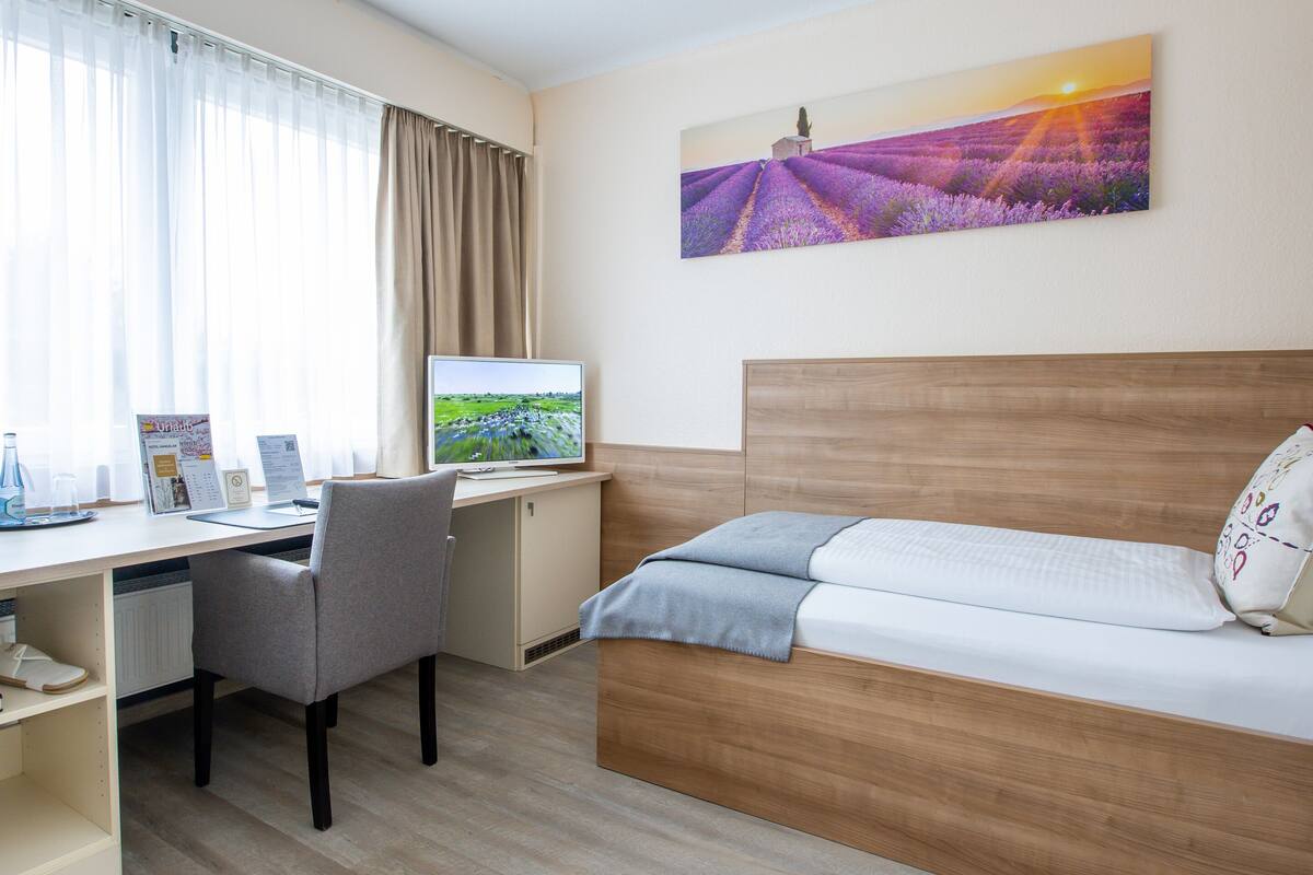 A hotel room with a wooden desk, a computer, a chair, a bed, and a large window with curtains.