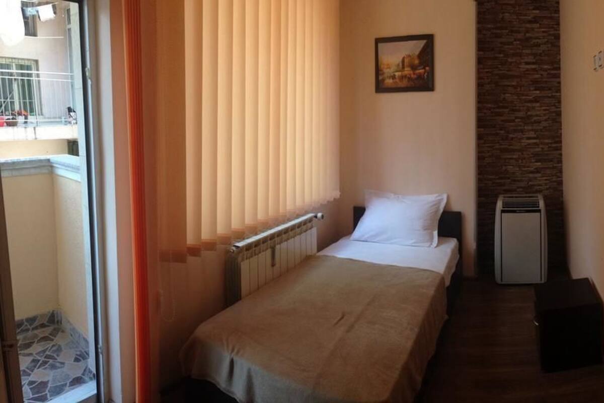 A small, neatly arranged bedroom with a single bed, a radiator, a small heater, and a door leading to an outdoor area.
