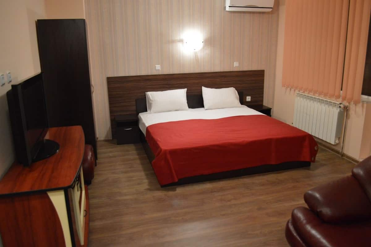 A hotel room with a wooden floor, a bed with a red bedspread, a TV on a wooden desk, and a radiator.