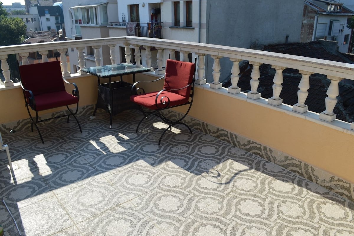 A balcony with patterned flooring, two red chairs, a small table, and a railing.