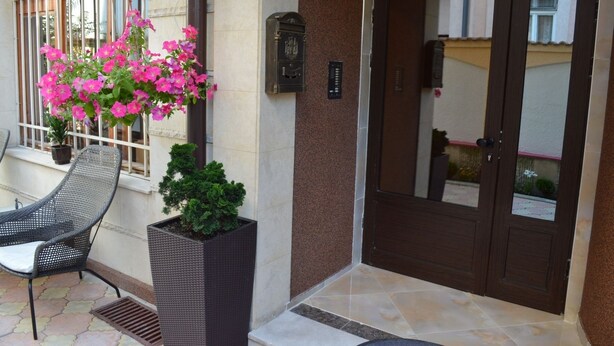 A hotel entrance with a dark wooden door, a potted plant with pink flowers, a wicker chair, and a small potted tree. - Bon Bon Hotel (Sofia)