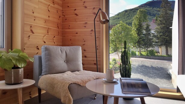 Ett mysigt rum med träväggar, en bekväm fåtölj, ett litet bord med en bok och en kopp, och utsikt över ett grönskande landskap med träd och en kulle.