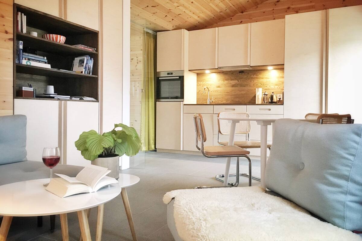 Een moderne woonkamer met een bank, een ronde tafel met een boek en een glas wijn, een boekenkast, en een keuken met een eettafel en stoelen.